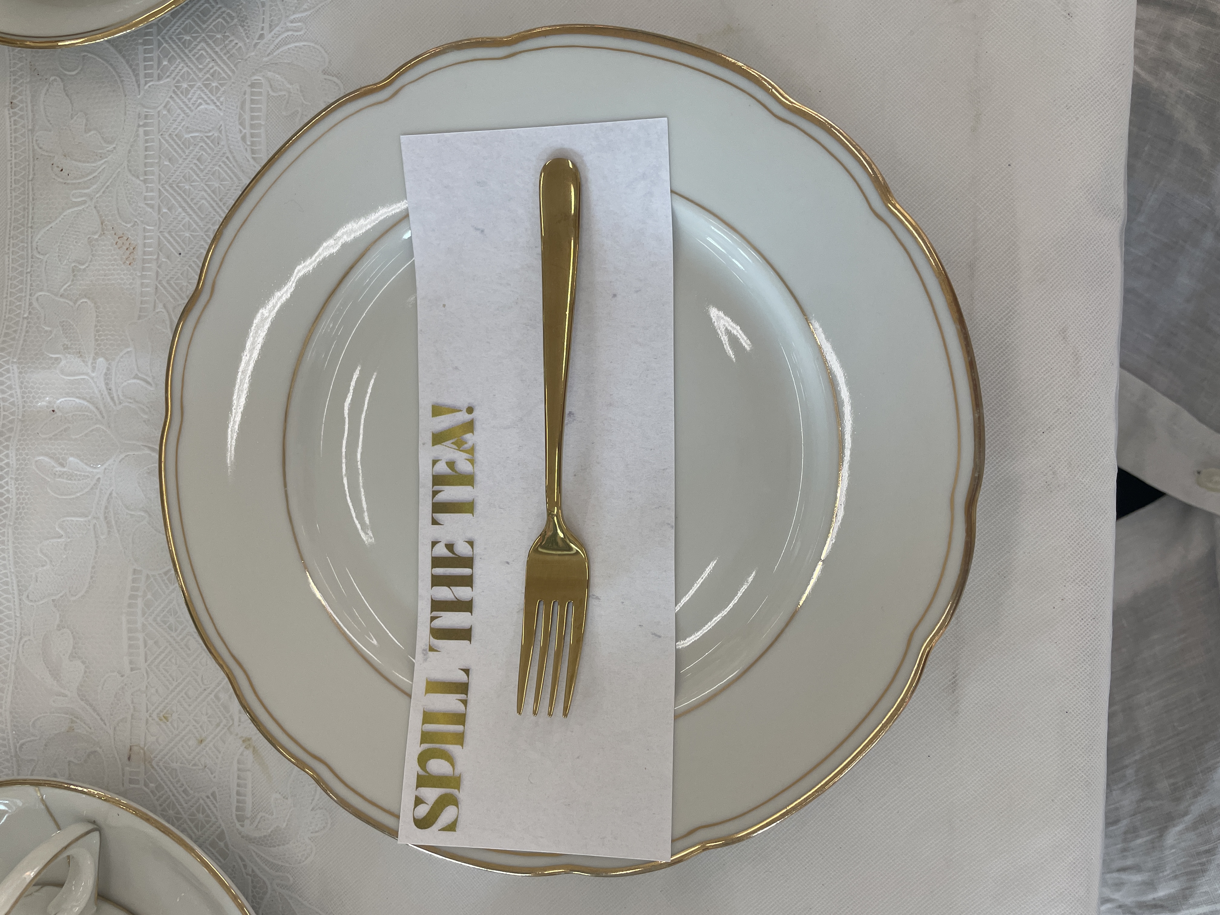 Ein Teller mti goldenem Rand. Darauf befindet sich eine goldene Gabel auf einer Serviette. Auf dieser steht: Spill the Tea in goldener Schrift