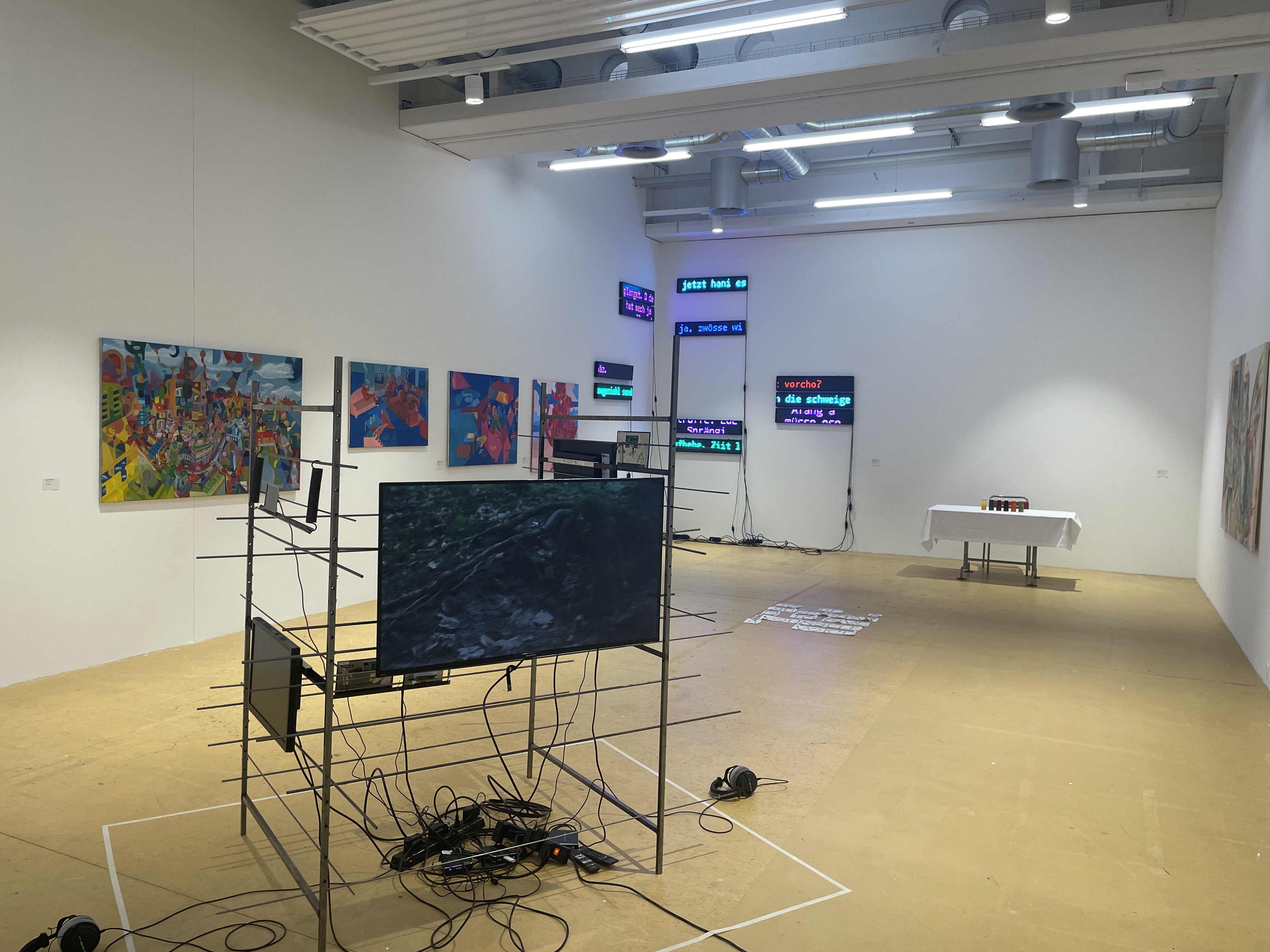 Ausstellungsraum mit einer Metallkonstruktion im Vordergrund. Im Hintergrund sind Malereien und Neon-Leuchtschilder