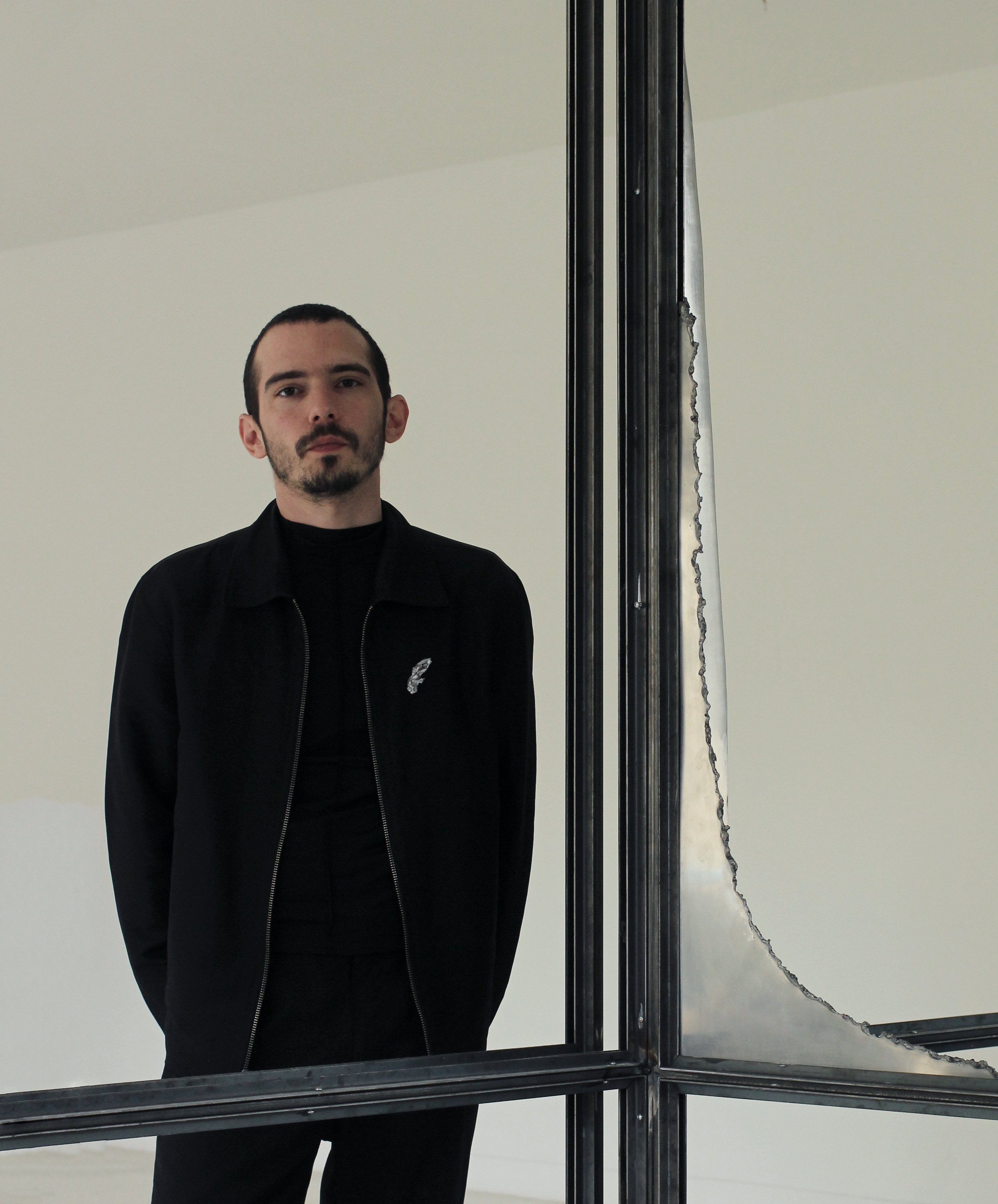 Portrait von Dimitris Tampakis. Der Künstler trägt schwarze Kleidung und blickt durch eine Metallkonstruktion. Im Hintergrund ist eine weisse Wand.