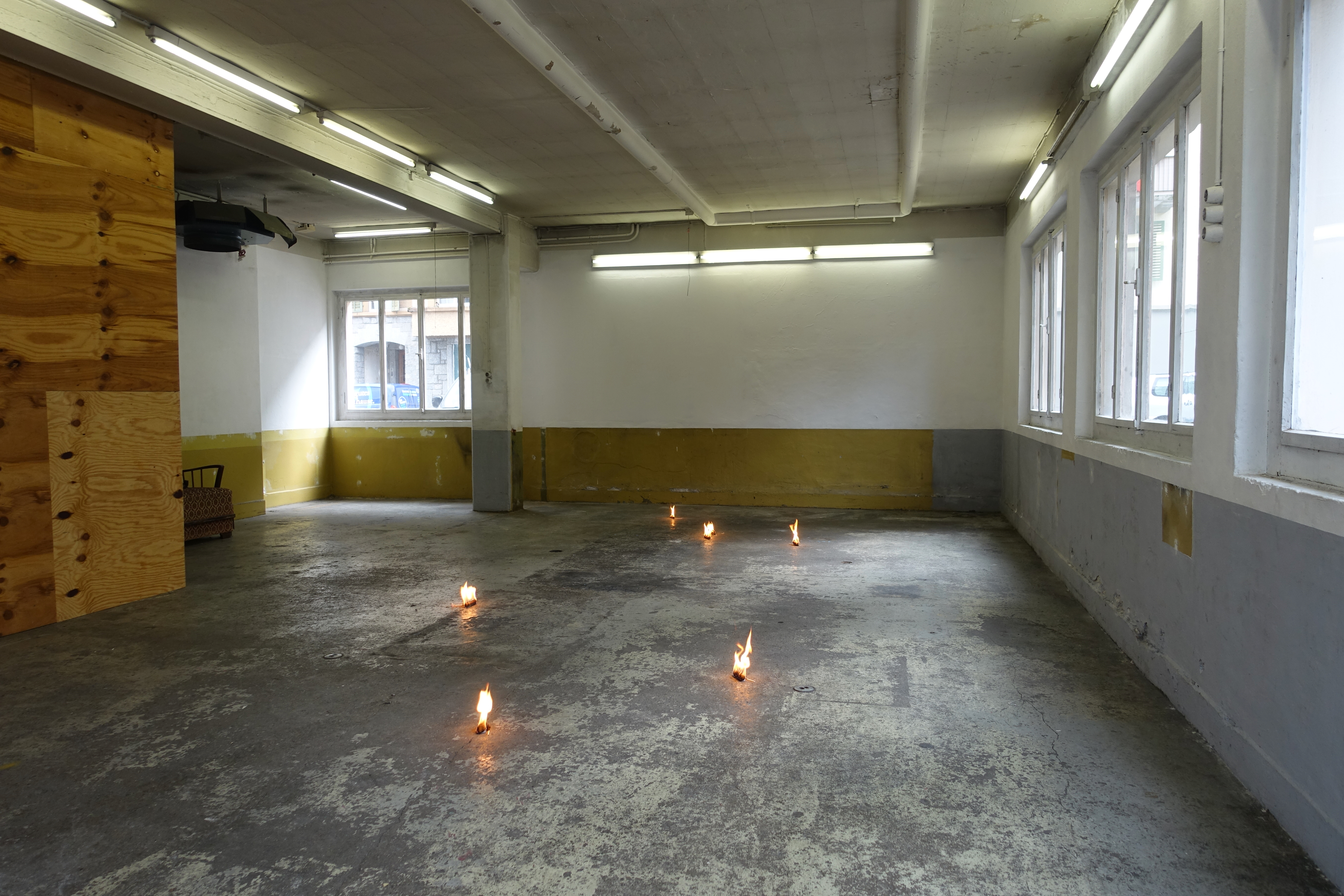 Ein Ausstellungsraum mit 6 brennenden kleinen Objekten auf dem Boden. Die Flammen sind nicht besonders hoch.