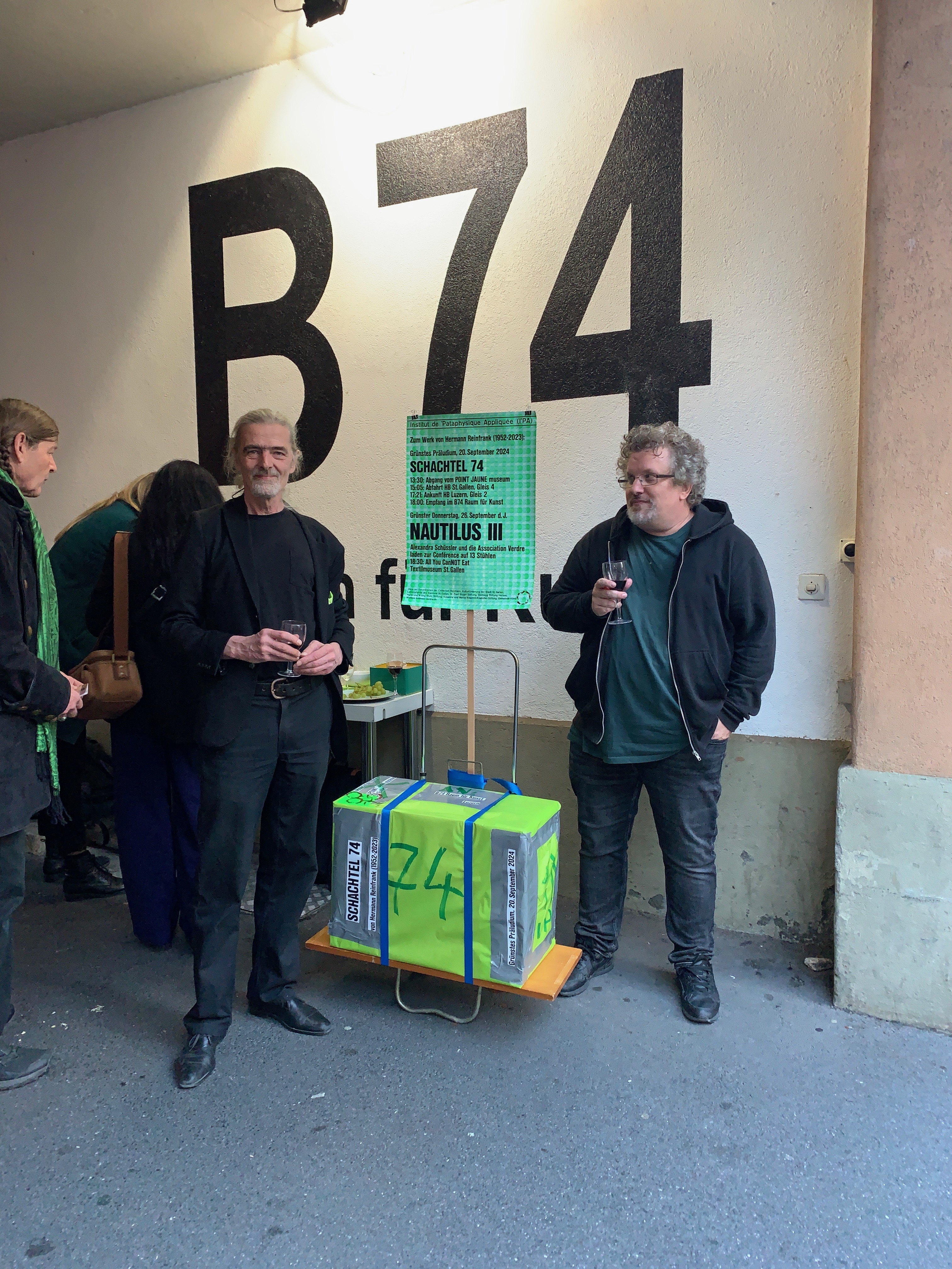 Zwei Personen stehen vor dem Eingang der Galerie B74 und halten Gläser in der Hand. Zwischen ihnen befindet sich eine leuchtend grüne Kiste.