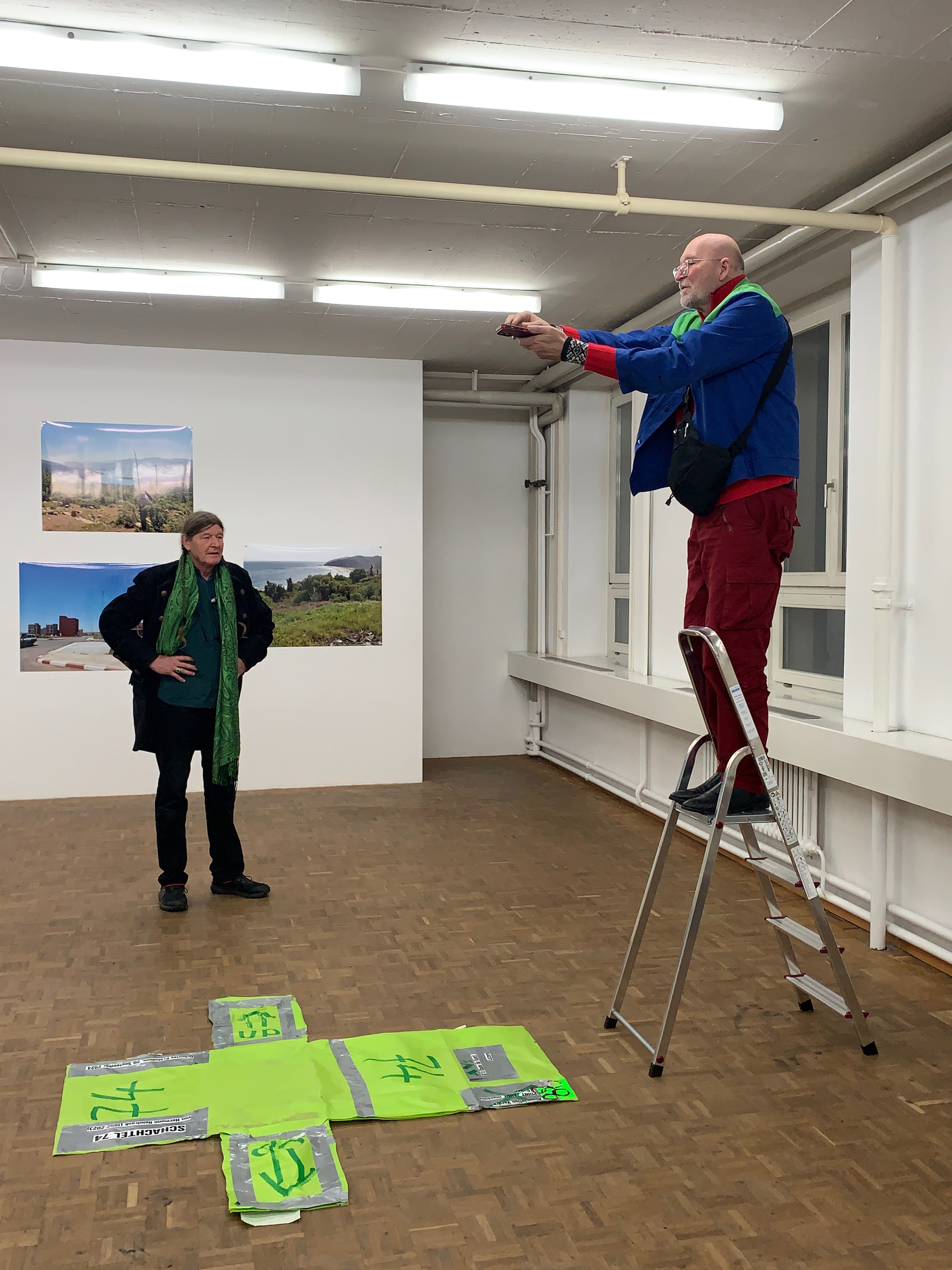 Auf einer Leiter befindet sich eine männlich gelesene Person. Sie fotografiert eine am Boden liegende Installation. im Hintergrudn ist eine weitere männlich gelesene Person zu erkennen. An der Wand befinden sich Bilder. Die Szenerie spielt in der Galerie.