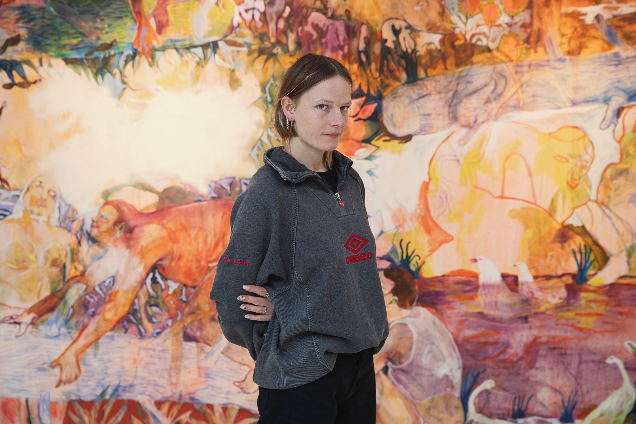 Portrait von Jeanne Jacob. Die Künstlerin steht vor einem farbigen Gemälde und trägt einen grauen Pullover. 