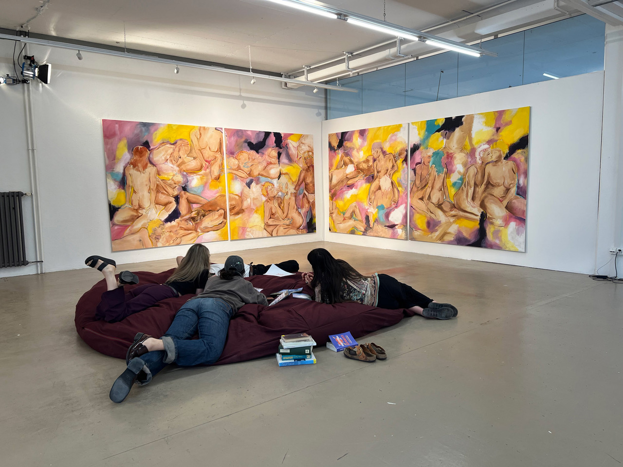 Eine Gruppe junger Menschen liegen auf einer Art Sitzsack auf dem Boden. Vor ihnen an der Wand sind 4 bunte Bilder zu sehen.