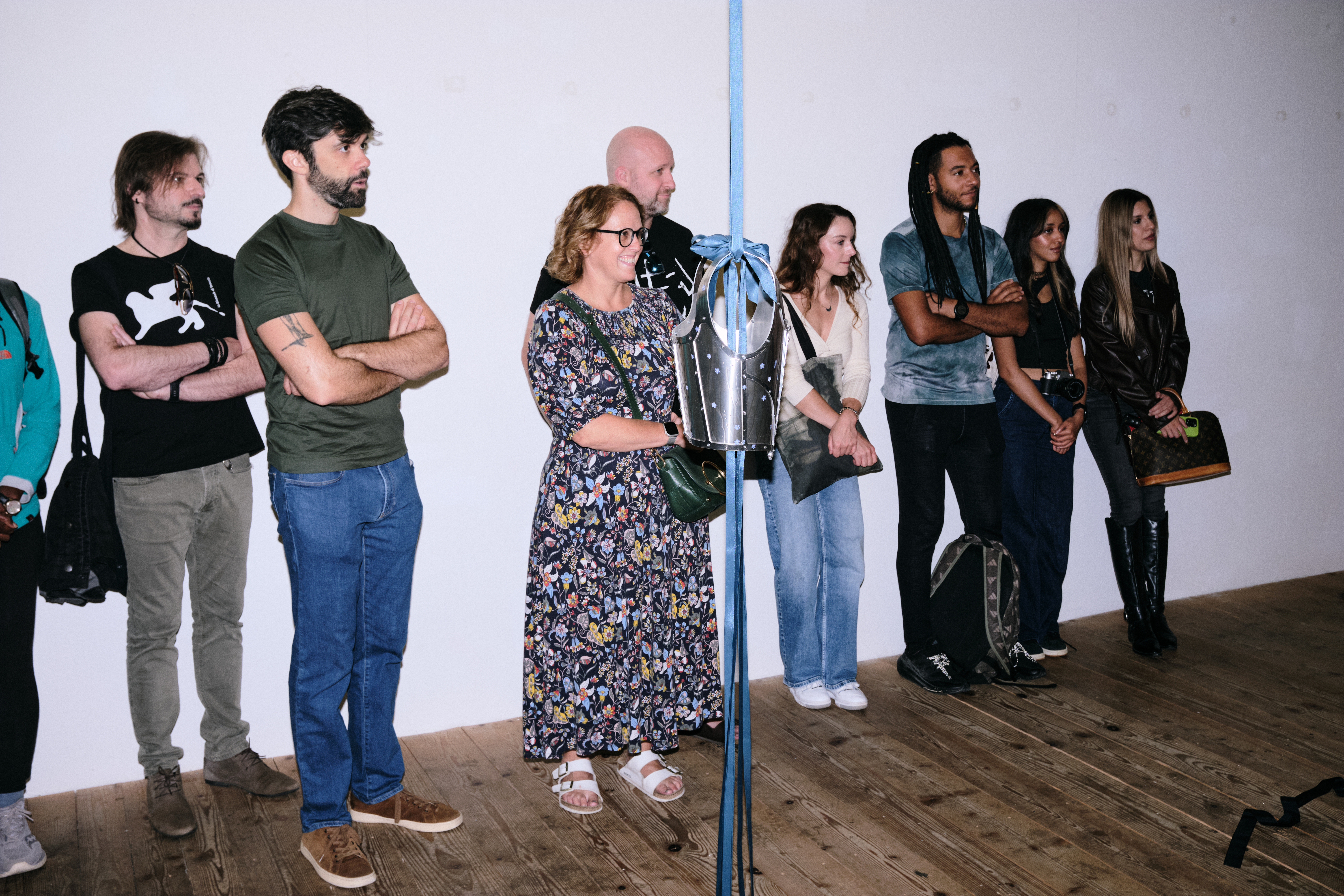 Foto von Shiva Gisiger Eine Gruppe steht im Ausstellungsraum. Von der Decke hängt eine Ritterrüstung an einem blauen Satinband