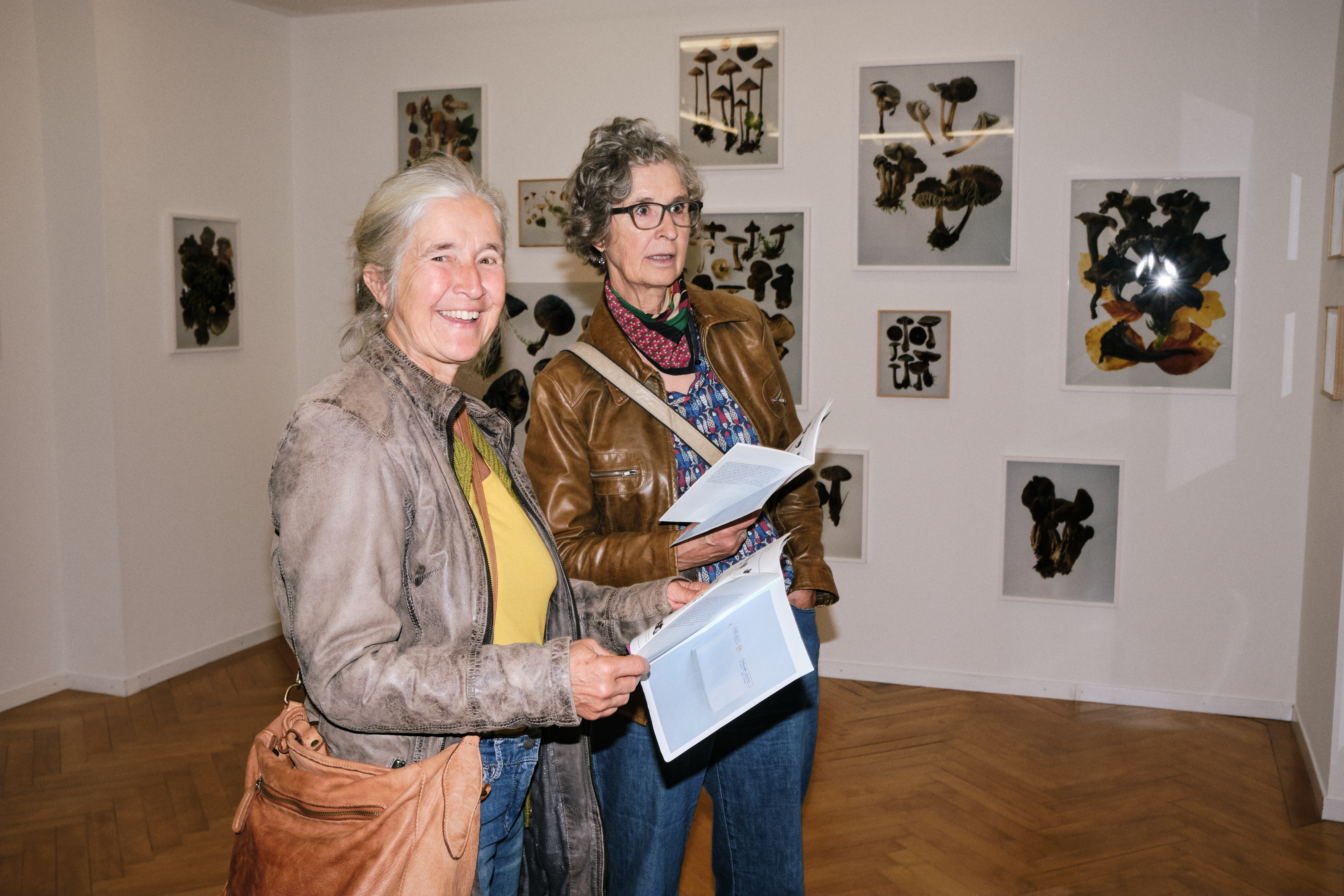 Foto von Shiva Gisiger Ausstellungsbesucherinnen blicken lachend in die Kamera. Im Hintergrund hängen Fotografien von Pilzen