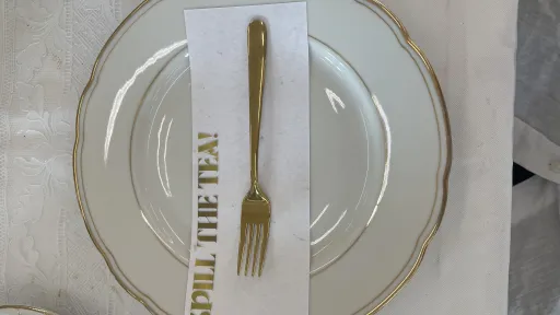 Ein Teller mti goldenem Rand. Darauf befindet sich eine goldene Gabel auf einer Serviette. Auf dieser steht: Spill the Tea in goldener Schrift
