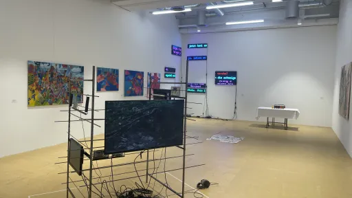 Ausstellungsraum mit einer Metallkonstruktion im Vordergrund. Im Hintergrund sind Malereien und Neon-Leuchtschilder