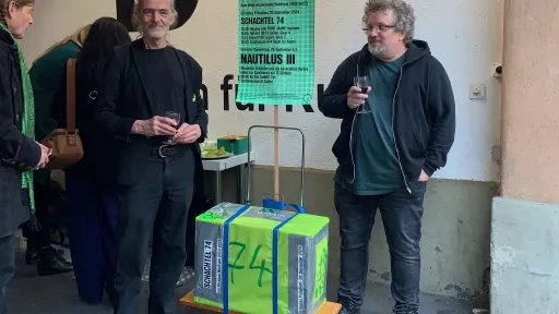 Zwei Personen stehen vor dem Eingang der Galerie B74 und halten Gläser in der Hand. Zwischen ihnen befindet sich eine leuchtend grüne Kiste.