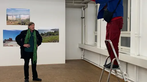 Auf einer Leiter befindet sich eine männlich gelesene Person. Sie fotografiert eine am Boden liegende Installation. im Hintergrudn ist eine weitere männlich gelesene Person zu erkennen. An der Wand befinden sich Bilder. Die Szenerie spielt in der Galerie.