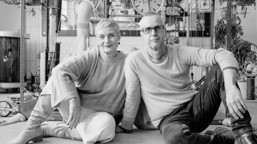 Eine Frau un ein Mann sitzen auf dem Boden vor einem Regal. Sie schauen beide in die Kamera