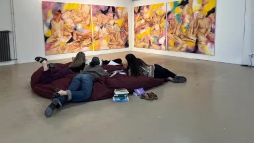 Eine Gruppe junger Menschen liegen auf einer Art Sitzsack auf dem Boden. Vor ihnen an der Wand sind 4 bunte Bilder zu sehen.