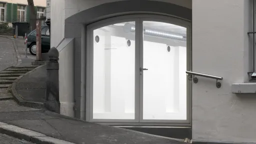 Eine gläserne Tür. Man sieht dahinter einen Ausstellungsraum. Die Tür hat einen leichten Bogen.