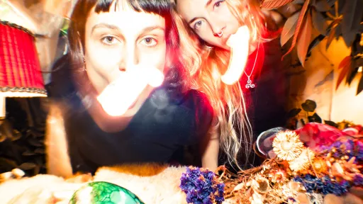 Laura Jana Luterbach & Naira Ramos, 2025 Foto : Leon Maria Plecity - Die zwei Künstlerinnen stehen vor einem Tisch mit bunten Objekten und Blumen